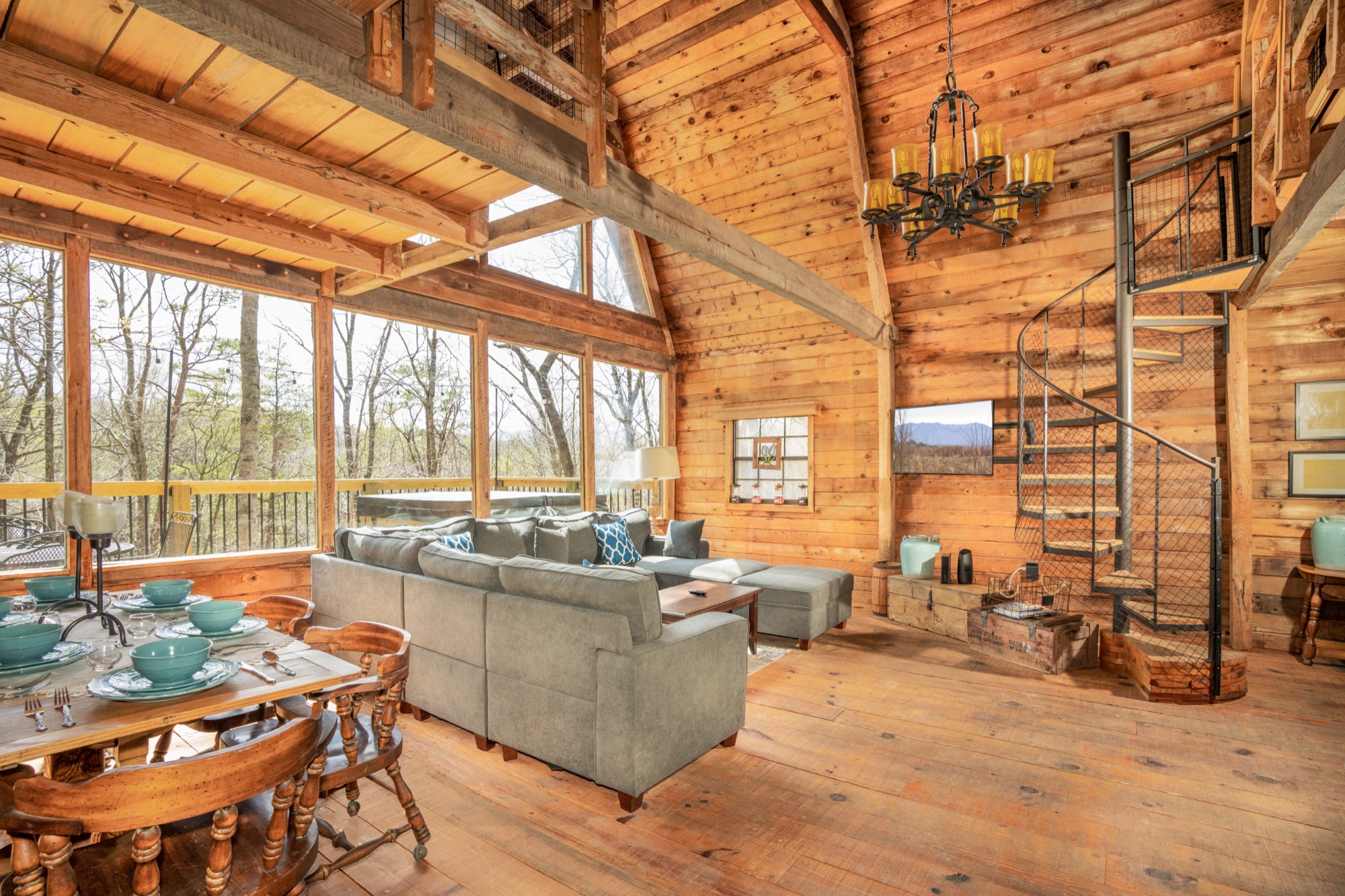 Rustic Retreat's great room with a spiral staircase, dining table, and sectional sofa beneath a soaring wood ceiling